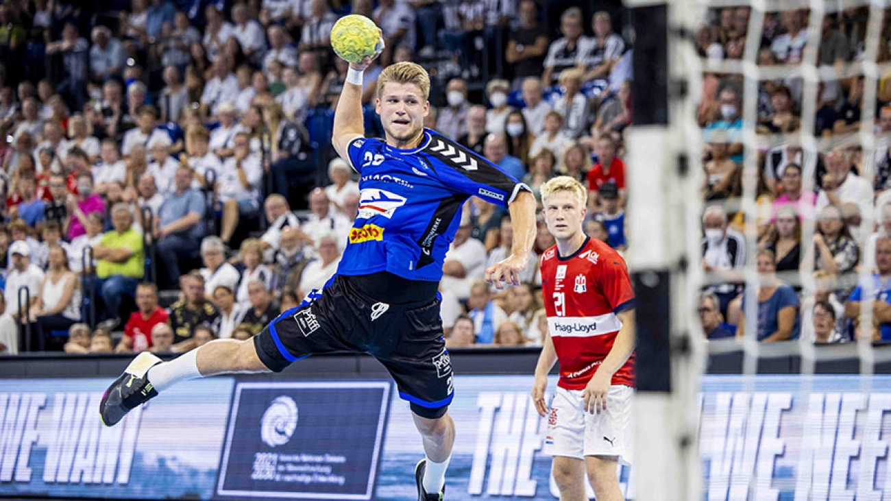 Ein Spieler des VfL Lübeck-Schwartau in blau-schwarzer Spielkleidung setzt mit einem gelben Handball zum Schuss an. Im Vordergrund sieht man die Maschen des Tors, im Hintergrund eine vollbesetzte Halle und einen gegnerischen Spieler in roter Spielkleidung