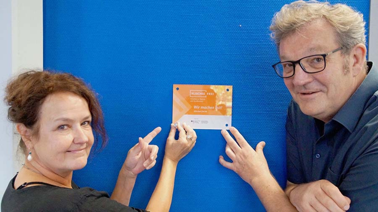 Eine Frau und ein Mann deuten vor einer blauen Wand auf einen orangen Zettel mit der Aufschrift "Klischeefrei"