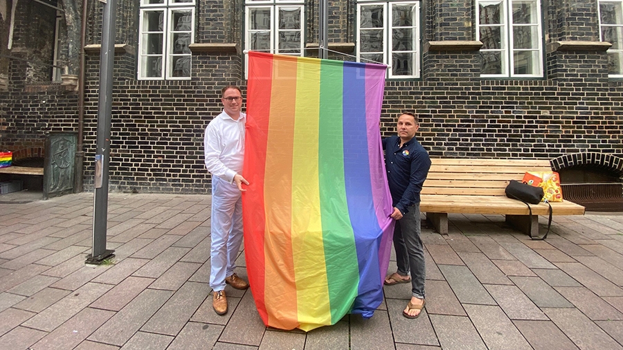 Zwei Männer vor einem dunklen Gebäude halten eine Regenbogenflagge fest