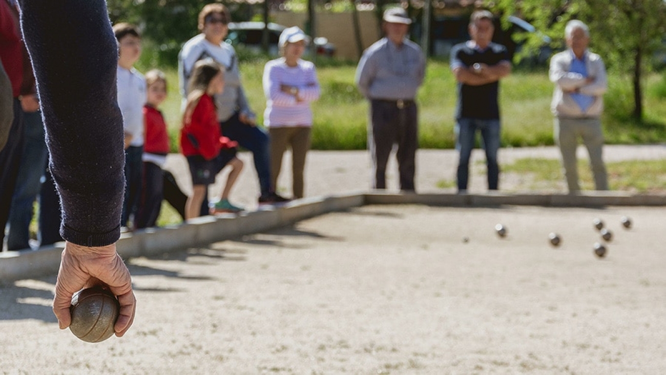 Eine Boulebahn mit Zuschauern, im Vordergrund sieht man eine Hand mit Boulekugel