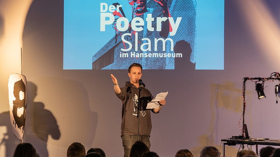 Auf einer Bühne steht ein Mann mit einem Blatt Papier in der Hand und spricht in ein Mikrofon. Im Hintergrund steht "Der Poetry Slam im Hansemuseum"