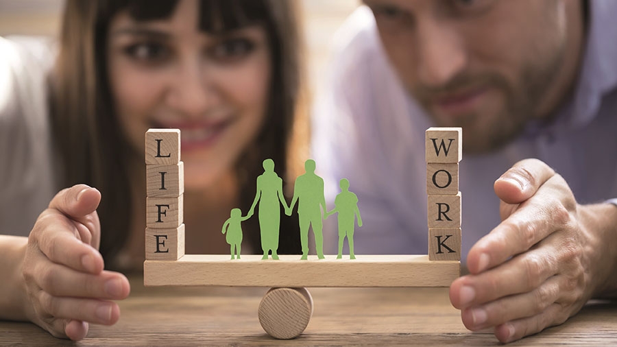 Ein Mann und eine Frau im Hintergrund halten eine Konstruktion im Vordergrund fest, auf der eine Familie und die Worte WORK und LIFE balancieren