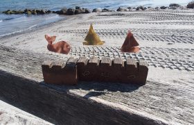 Auf einem Holzbalken an einem Strand steht ein bearbeitetes Stück Holz mit aufgesteckten Kupfer-Fischen und -Schiffen