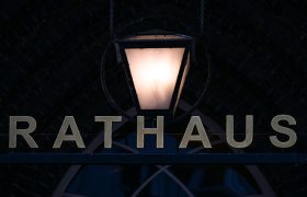 Über einem metallenen Schriftzug "Rathaus" leuchtet eine einzelne Laterne. Das Bild ist insgesamt sehr dunkel.