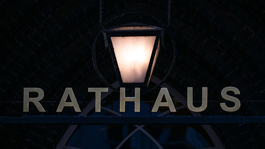 Über einem metallenen Schriftzug "Rathaus" leuchtet eine einzelne Laterne. Das Bild ist insgesamt sehr dunkel.