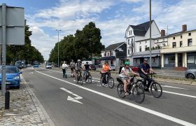 Eine Gruppe Fahrradfahrer ist im urbanen Raum auf einer Straße unterwegs. Rechts davon ist eine Rechtsabbiegerspur frei.