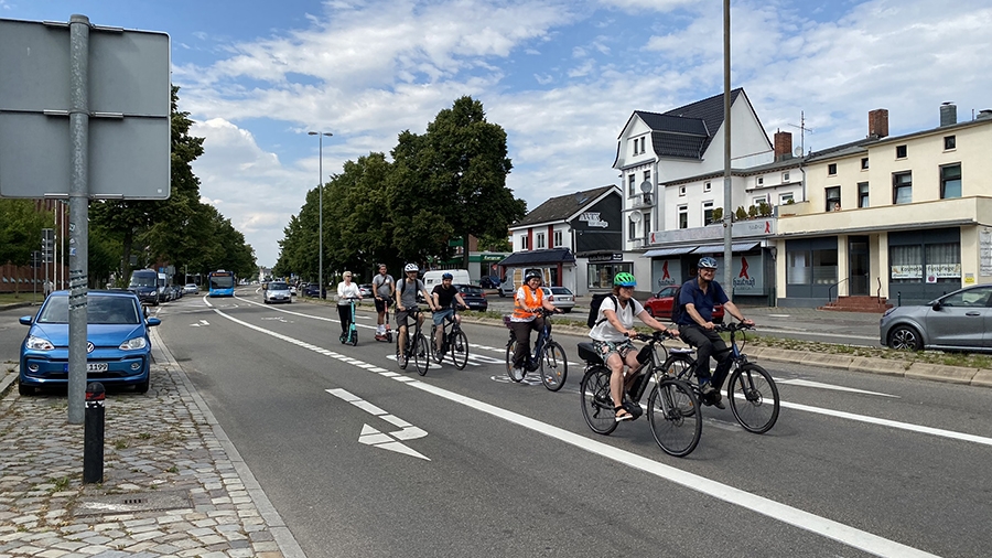 Eine Gruppe Fahrradfahrer ist im urbanen Raum auf einer Straße unterwegs. Rechts davon ist eine Rechtsabbiegerspur frei.