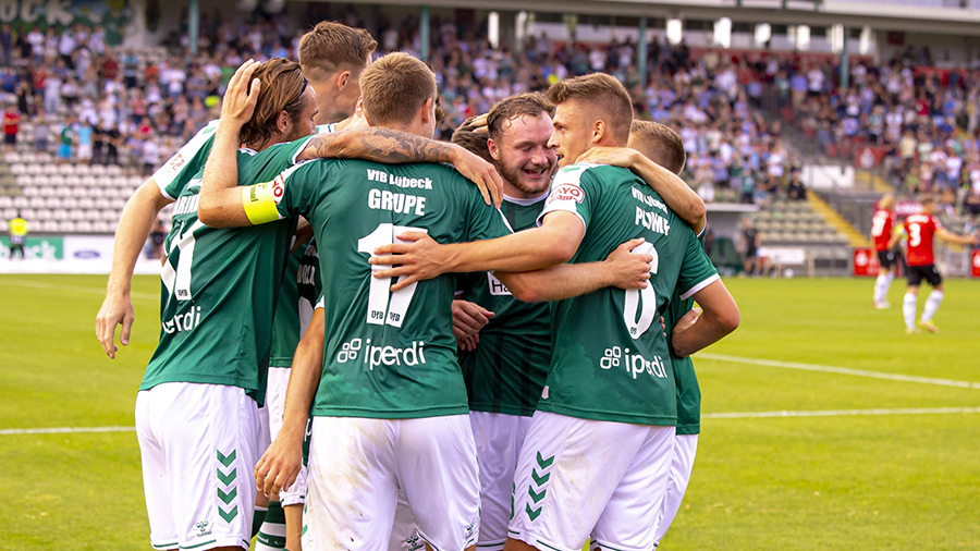 Eine Gruppe Fußballspieler des VfB Lübeck mit grünen Trikots und weißen Hosen umarmt sich auf einem Fußballplatz