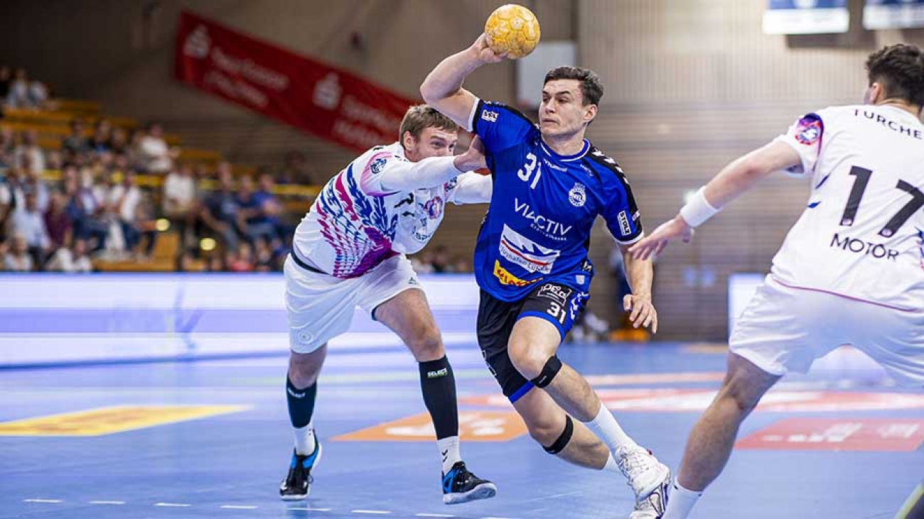Im Zentrum des Bildes sieht man einen Spieler des VfL Lübeck-Schwartau in blauer Spielkeidung, der mit einem gelben Handball zum Schuss ansetzt. Zwei Spieler in weißer Spielkleidung attackieren ihn