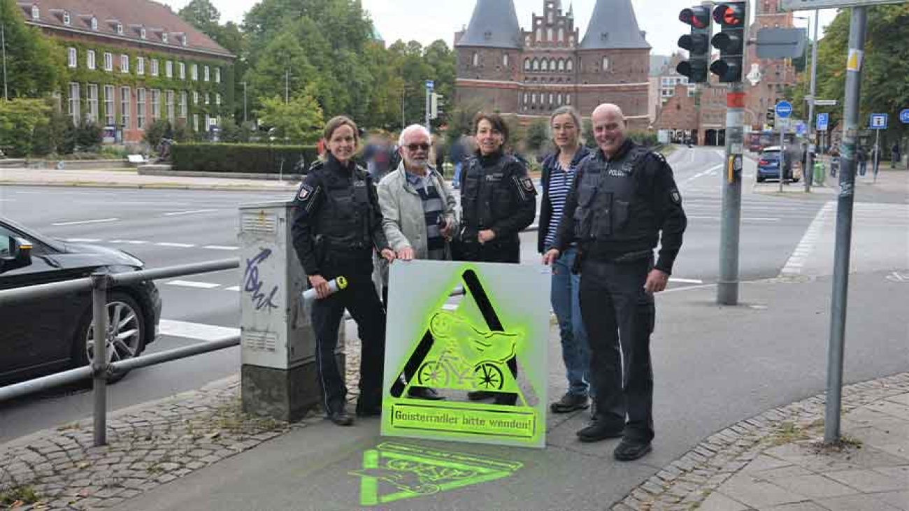 Vor dem Holstentor steht eine Gruppe Menschen, darunter eine Frau und ein Mann in Polizeiuniform, Sie halten eine Schablone, mit der auf dem Radweg in neongelb ein Verkehrszeichen und die Aufschrift "Geisterradler, bitte wenden!" gesprüht wurde