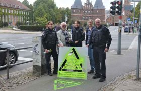 Vor dem Holstentor steht eine Gruppe Menschen, darunter eine Frau und ein Mann in Polizeiuniform, Sie halten eine Schablone, mit der auf dem Radweg in neongelb ein Verkehrszeichen und die Aufschrift "Geisterradler, bitte wenden!" gesprüht wurde