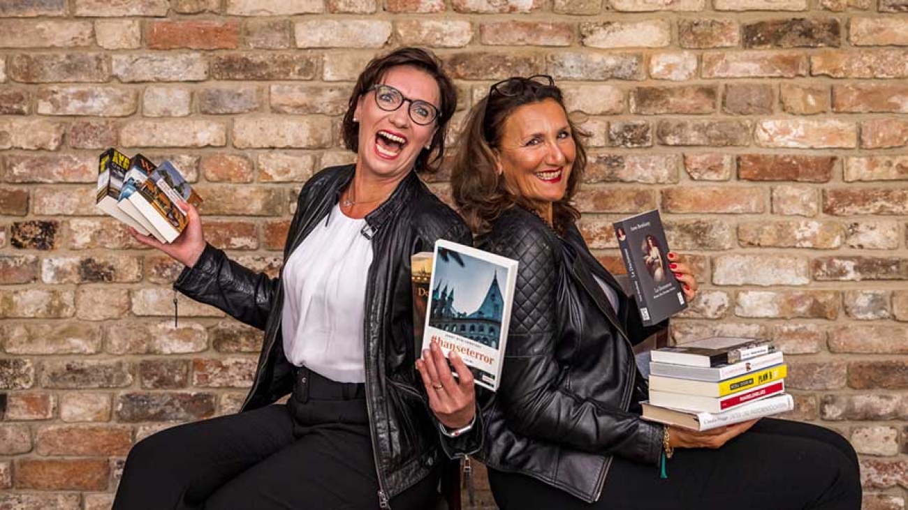 Vor einer Backsteinmauer sitzen zwei fröhliche Frauen in schwarzen Lederjacken und halten Bücher in den Händen