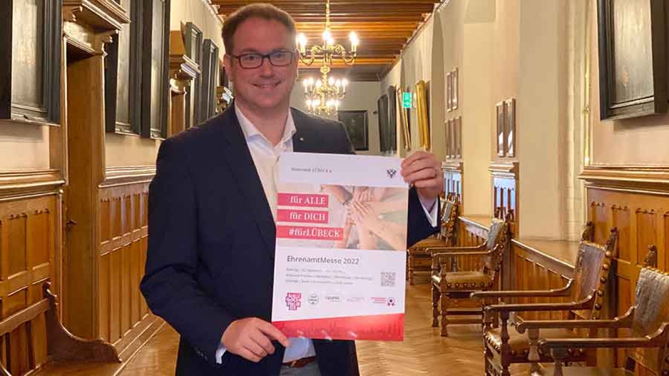 Lübecks Bürgermeister Jan Lindenau zeigt in einem repräsentativen Flur des Rathauses ein Plakat der Ehrenamtmesse in die Kamera