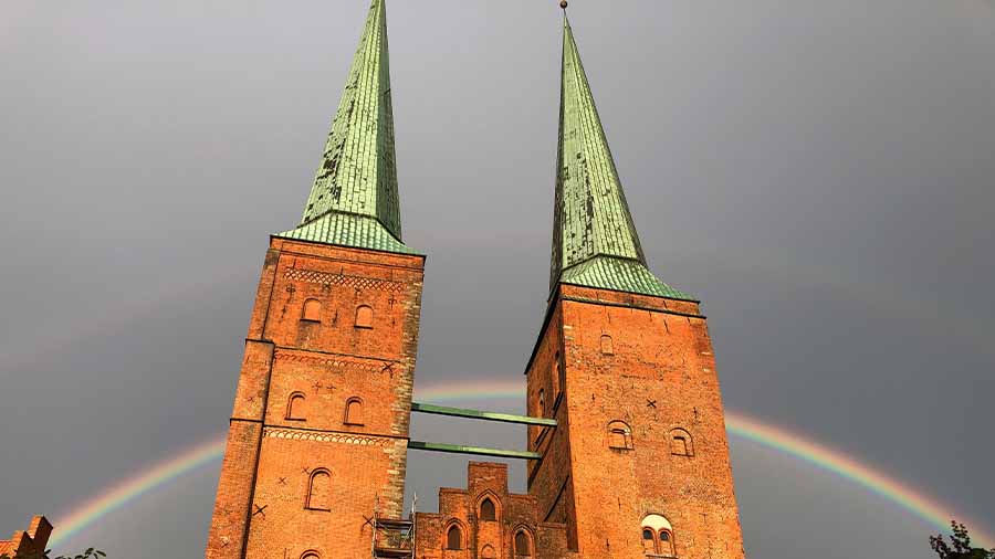 Vor einem grauen Himmel mit einem Regenbogen sieht man die angeleuchteten Türme des Lübecker Doms