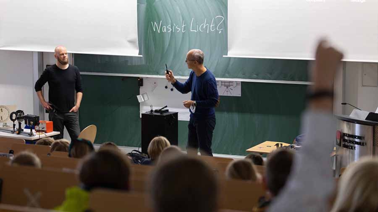 Blick von den Rängen in einen Hörsaal. Zwei Personen stehen vor einer Tafel, auf der "Was ist Licht?" steht.