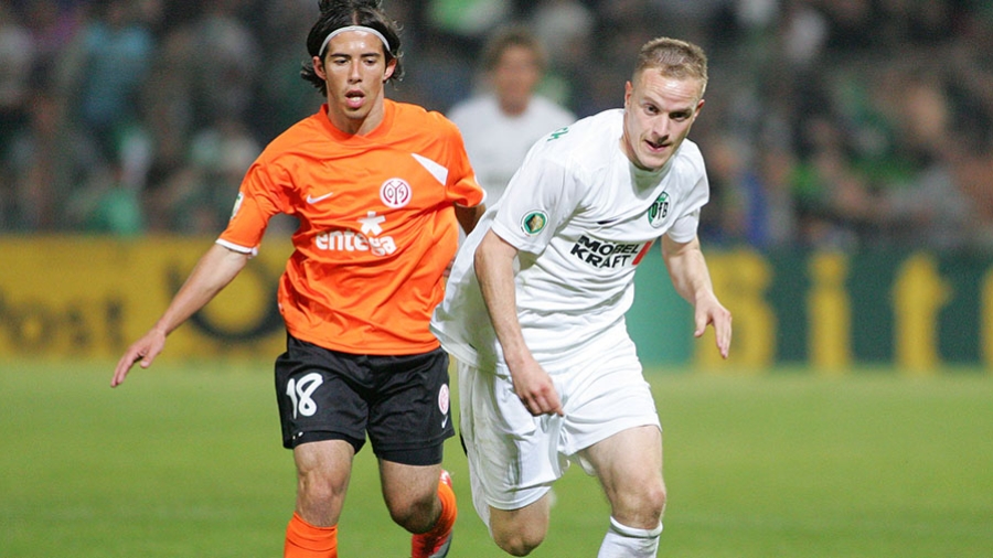 Ein Spieler des VfB Lübeck in weißer Spielkleidung läuft neben einem Spieler des FSV Mainz 05 in orange-schwarzer Spielkleidung her