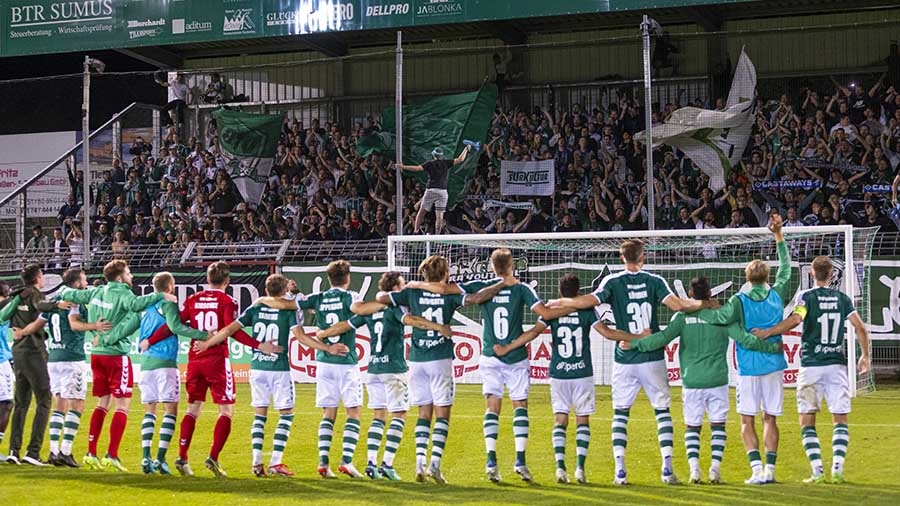 Eine Reihe Fußballspieler des VfB Lübeck in grünen Trikots und weißen Hosen steht vor der Fankurve und jubelt