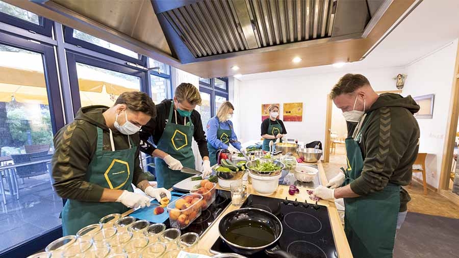 Mehrere Männer des VfB Lübeck in olivgrünen Trainingsjacken und grünen Schürzen bereiten an einer Kochinsel ein Essen vor