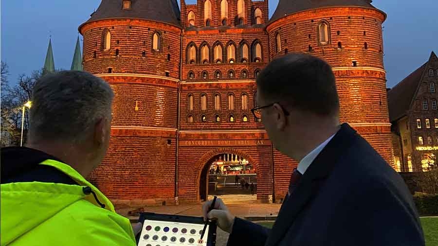 Vor dem abendlich angestrahlten Holstentor stehen zwei Männer mit einem digitalen Endgerät und regeln die Lichtfarbe