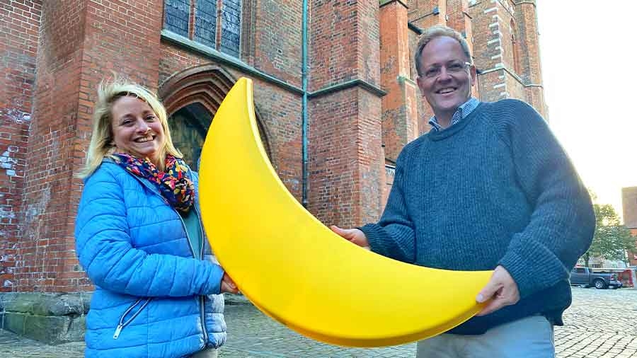 Zwei Menschen halten vor einer gotischen Backsteinfassade eine große gelbe Mondsichel