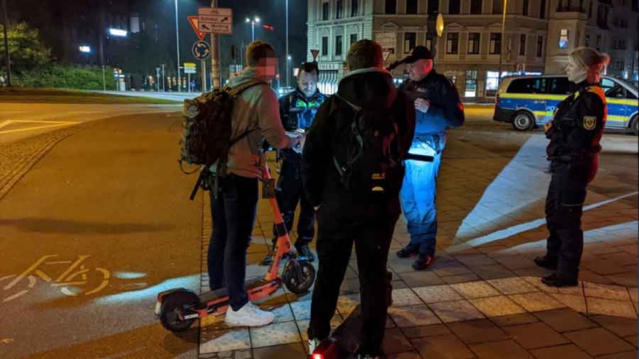 In einer abendlichen Szene stehen mehrere Polizisten und zwei Menschen auf E-Scootern beisammen. Die Polizisten kontrollieren die Scooter-Fahrer. Es ist Taschenlampenlicht zu sehen.