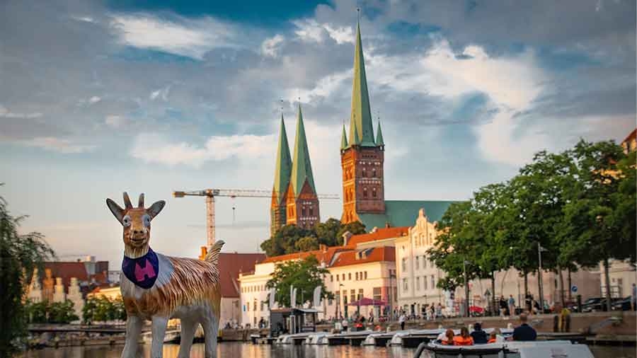 Vor der Stadtkulisse Lübecks steht eine lebensgroße Modellziege und schaut in die Kamera. Sie trägt ein Halstuch mit der Silhouette des Holstentors.