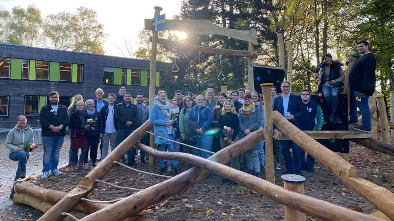 Im Vordergrund ein Klettergerüst aus Baumstämmen und Seilen, direkt dahinter ein Gruppenbild von rund 30 Menschen. Im Hintergrund ein Zweckbau aus Holz mit grünen Fensterläden und hoher Baumbestand