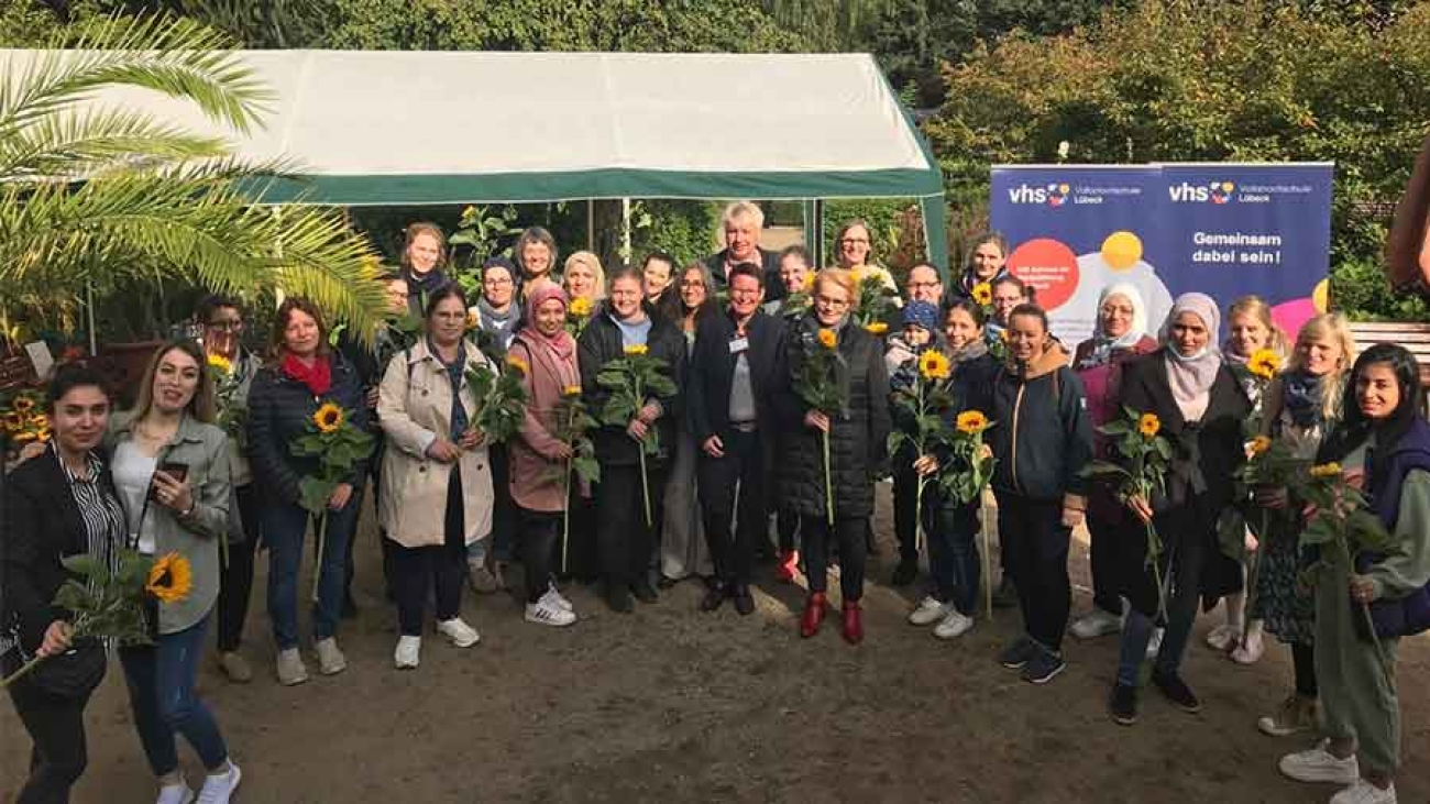 In einem Gartengelände stehen im Halbkreis eine Gruppe von Frauen mit Sonnenblumen in den Händen