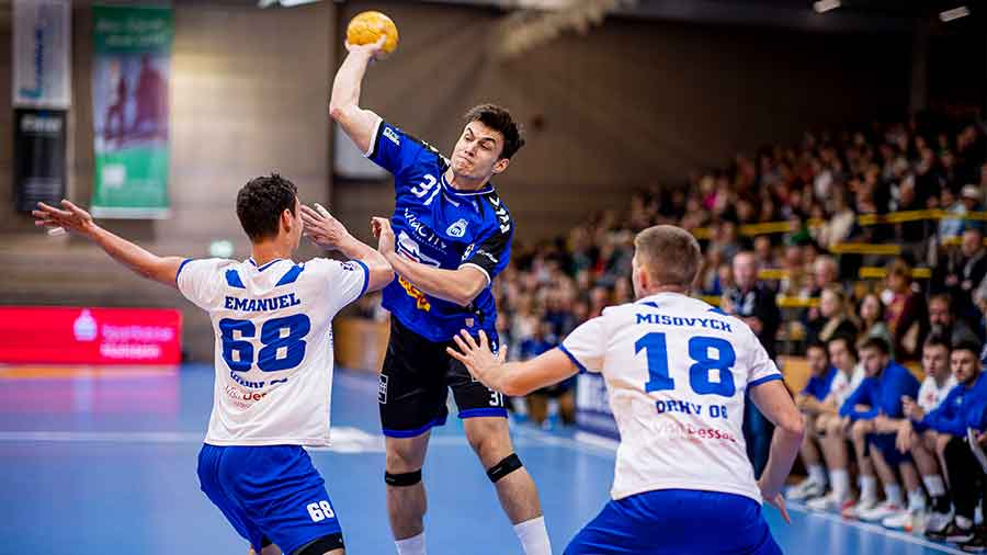 Der Handballer Matej Klima in blau-schwarzer Spielkleidung setzt zum Wurf an. Ihm gegenüber stehen zwei Spieler vom DRHV in weiß-blauer Spielkleidung
