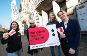Vier Menschen stehen mit dem Plakat "Das ganze Theater umsonst!" vor dem Theater Lübeck und zeigen Tickets in die Kamera