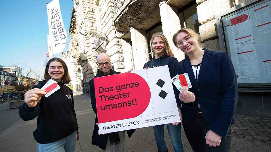 Vier Menschen stehen mit dem Plakat "Das ganze Theater umsonst!" vor dem Theater Lübeck und zeigen Tickets in die Kamera