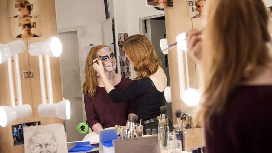 Ein Blick in einen Theaterspiegel: Eine junge Frau schminkt eine andere mit einem aufwändigen Theater-Make-Up