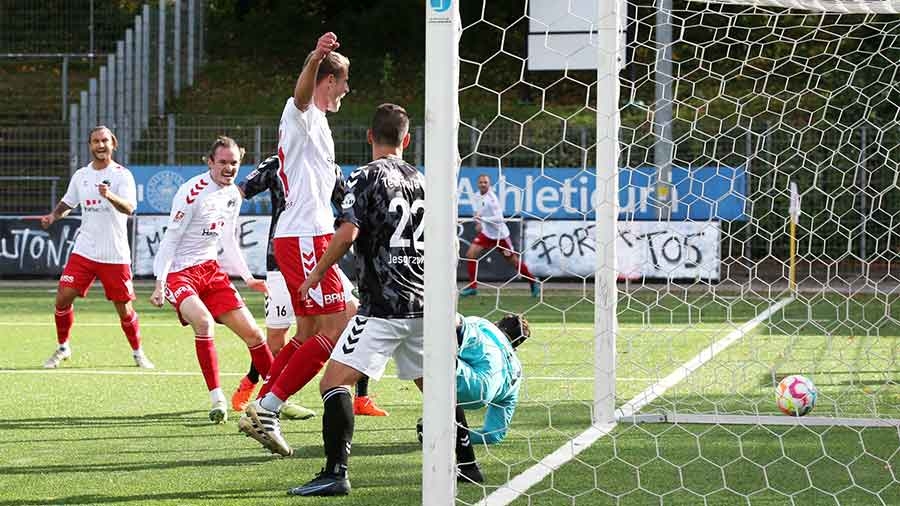 Vor einem Fußballtor jubeln mehrere Spieler des VfB Lübeck in weißen Auswärtstrikots und roten Hosen. Der Ball liegt im Tor