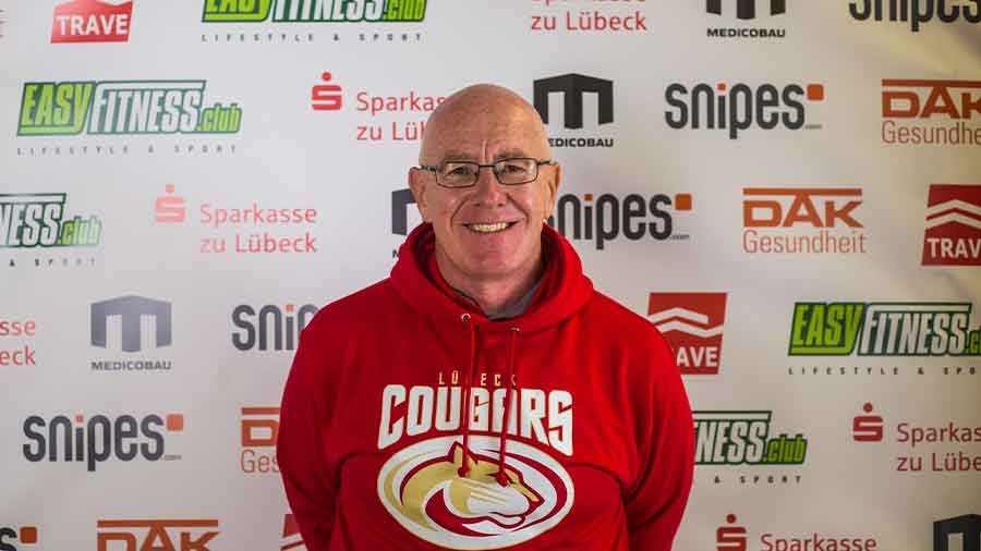 Ein Mann mit Halbglatze und kurzen Haaren in rotem Hoodie mit dem Logo der Lübeck Cougars lächelt freundlich in die Kamera