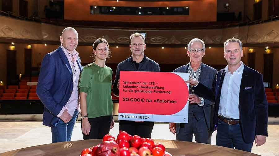 Eine Gruppe Menschen steht auf der Bühne eines Theaters. Im Hintergrund ist der Zuschauerraum zu sehen. Sie halten ein Schild mit der Aufschrift "Wir danken der LTS - Lübecker Theaterstiftung für die großzügige Förderung! 20 000 Euro für "Salome"