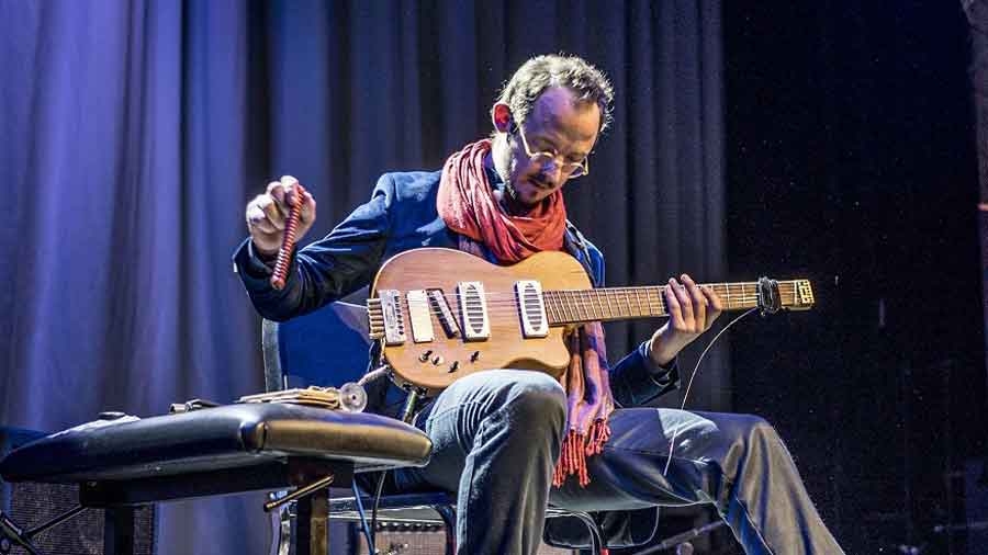 Auf einer Bühne sitzt ein Mann mit rotem Schal. Er hält eine E-Gitarre auf den Knien und einen Klangstab in der Hand.