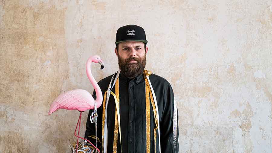 Der Künstler Patrick Salmen steht mit ernstem Gesicht vor einer beige Wand. In der Hand trägt er einen rosa Flamingo, er hat Luftschlangen um den Hals