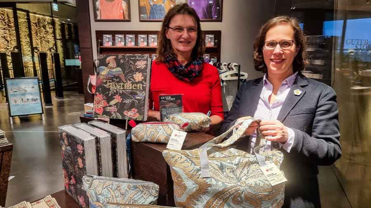 Zwei Frauen stehen in der Umgebung einer Ausstellung. Sie zeigen Taschen aus Brokatstoff