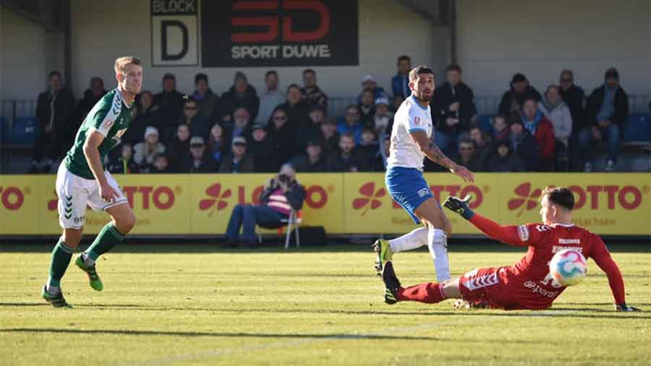 Auf einem sonnebeschienenen Fußballplatz schaut ein Spieler des VfB Lübeck in grünem Trikot und weißen Hosen einem Ball hinterher. Ein Spieler des SSV Jeddeloh in weißem Trikot und blauen Hosen schaut ebenfalls, ein Torwart in roter Sportkleidung liegt auf dem Boden