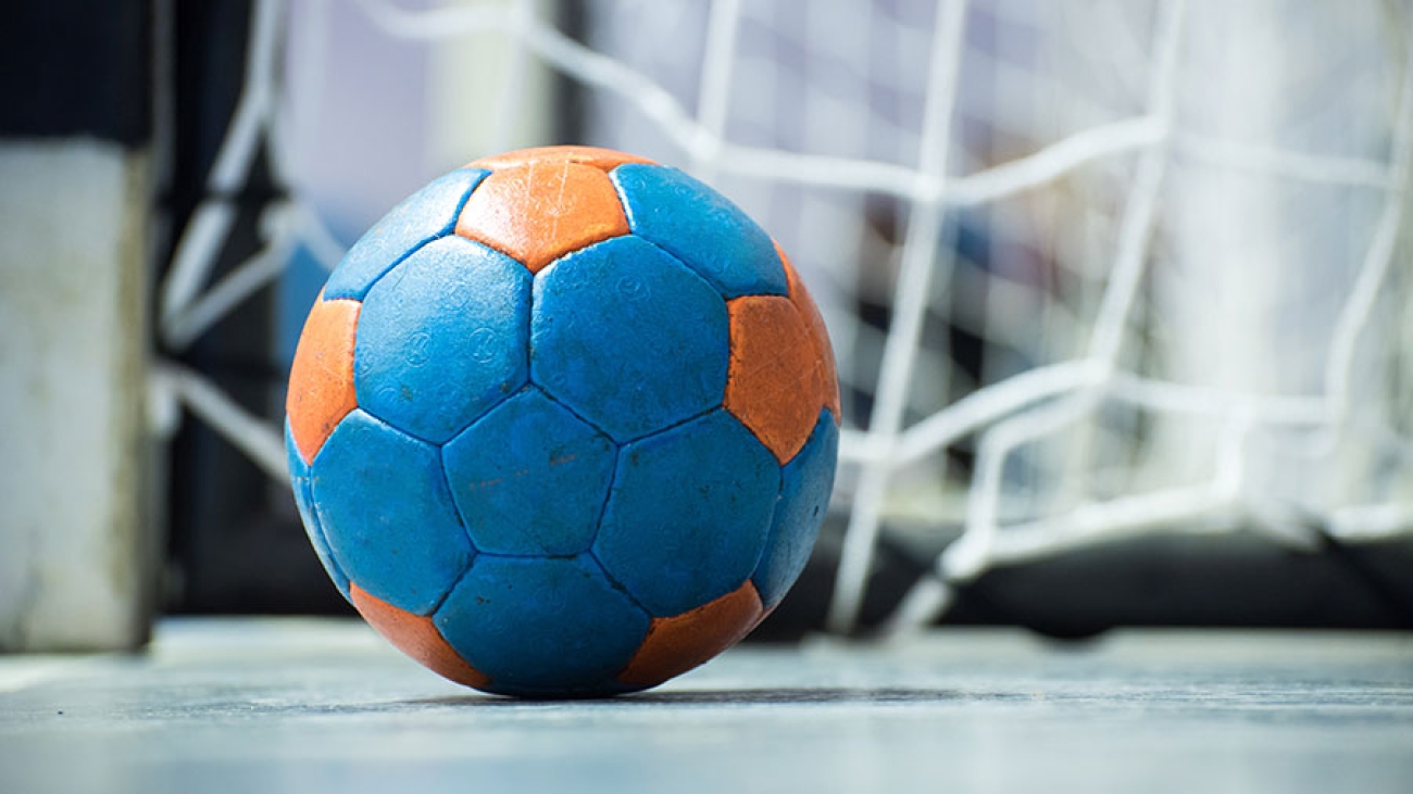 Handball in blau und orange liegt auf dem Boden vor einem Tor.