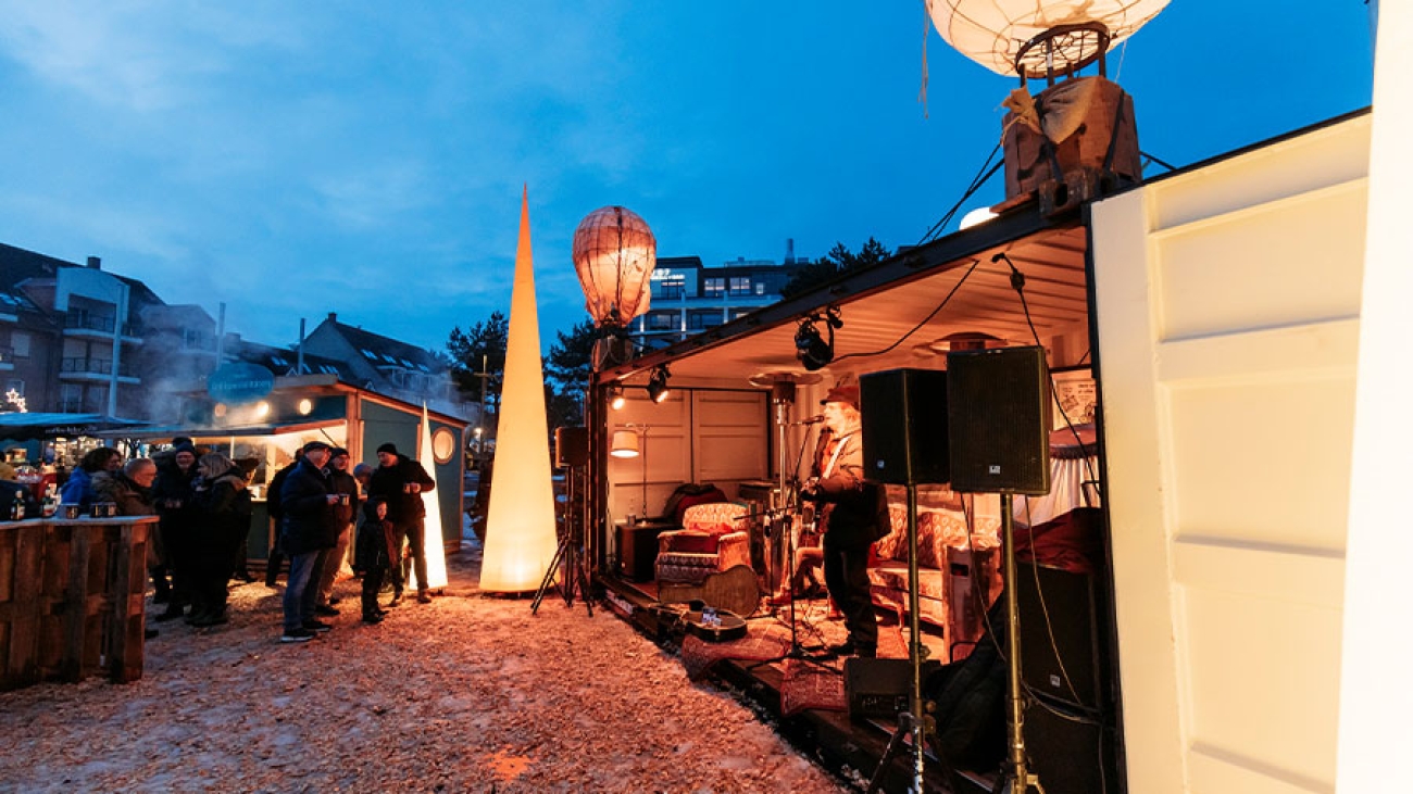 In einem geöffneten Container unter freiem Himmel spielt ein Mann Gitarre auf einer Wohnzimmerbühne. Mehrere Menschen stehen vor Imbissbuden und schauen zu. Auf dem Boden liegen Schnee und Holzschnitzel