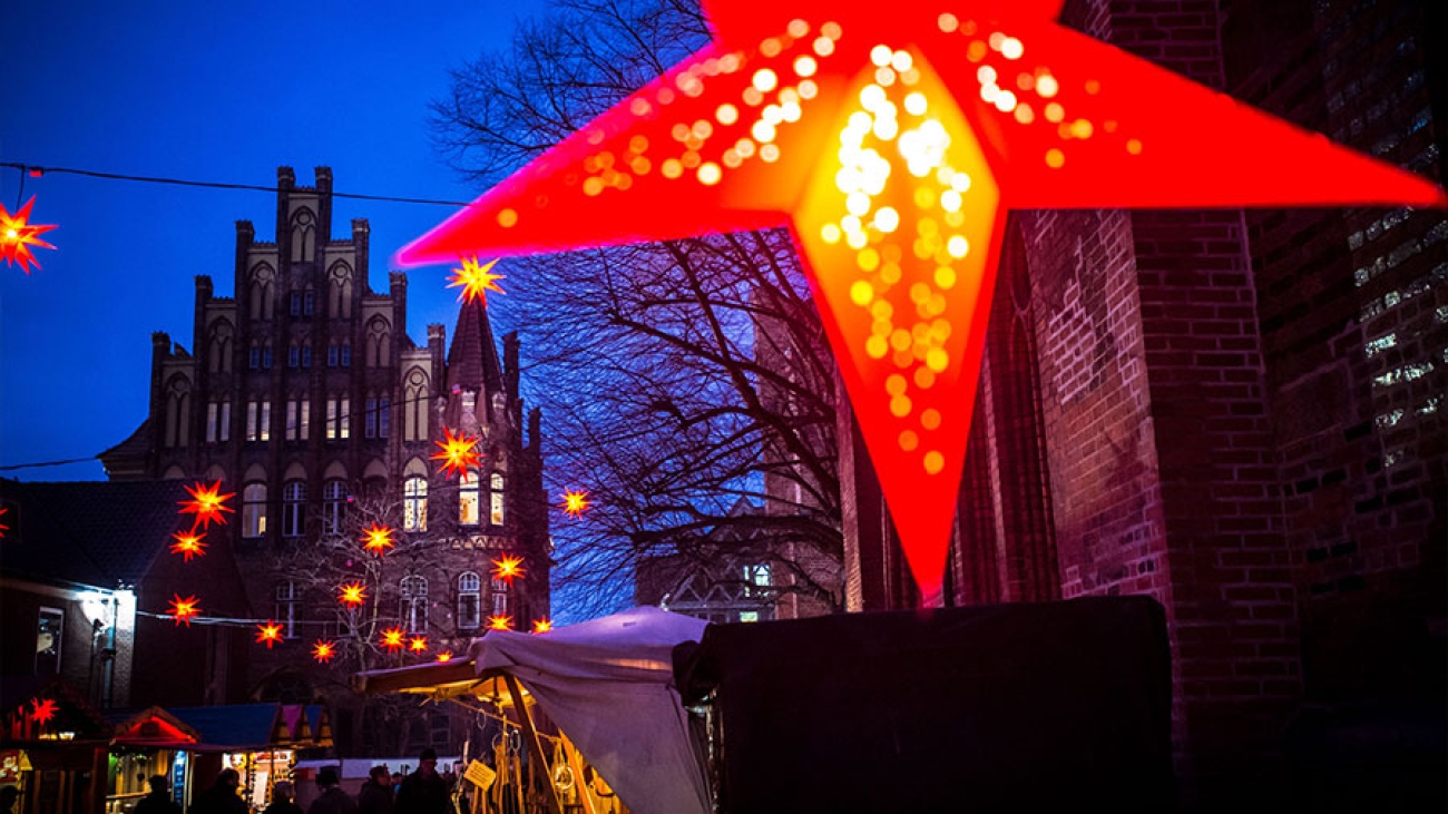 Hinter einem prominent im Bild hängenden rot beleuchteten Weihnachtsstern sieht man die Szenerie eines abendlichen Weihnachtmarktes mit roten Sternen in der Luft und einem gotischen Treppengiebel im Hintergrund