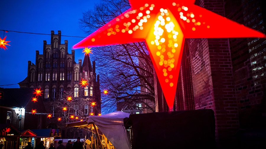 Hinter einem prominent im Bild hängenden rot beleuchteten Weihnachtsstern sieht man die Szenerie eines abendlichen Weihnachtmarktes mit roten Sternen in der Luft und einem gotischen Treppengiebel im Hintergrund
