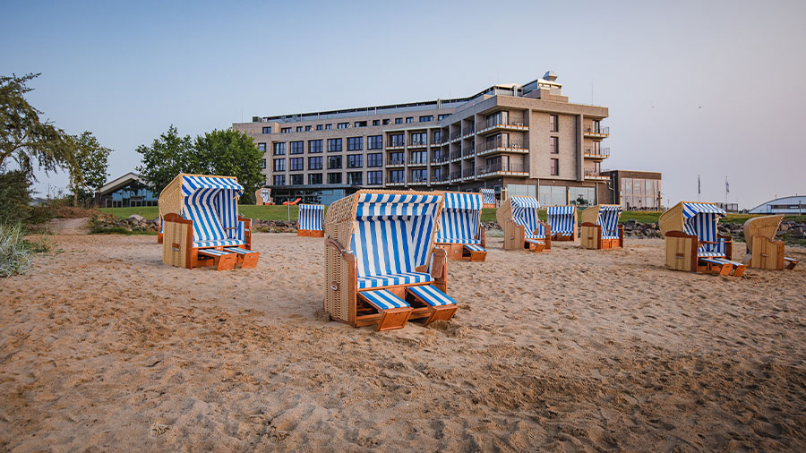 An einem Strand stehen vor einem Hotelbau mehrere blau-weiß gestreifte Strandkörbe