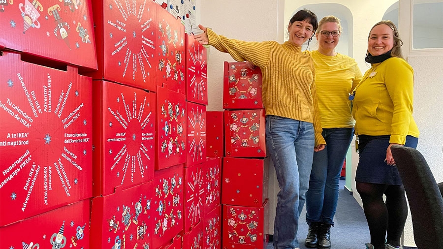 Drei Frauen mit gelben Pullovern stehen lachend neben roten Kartons mit weißer Aufschrift