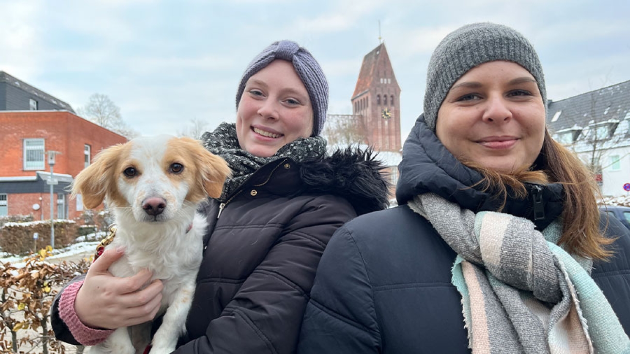 Zwei junge Frauen in Winterkleidung stehen im Freien. Im Hintergrund ein Kirchturm. Eine Frau hält einen weiß-braunen Hund im Arm.