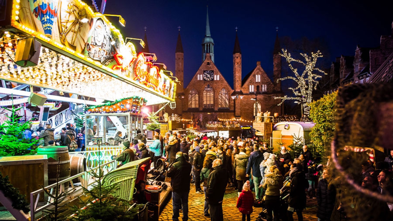 Ein Blick auf den Maritimen Weihnachtsmarkt. Links der beleuchtete Eingang zum Riesenrad, davor ein Strandkorb, im Hintergrund das Heilig-Geist-Hospital