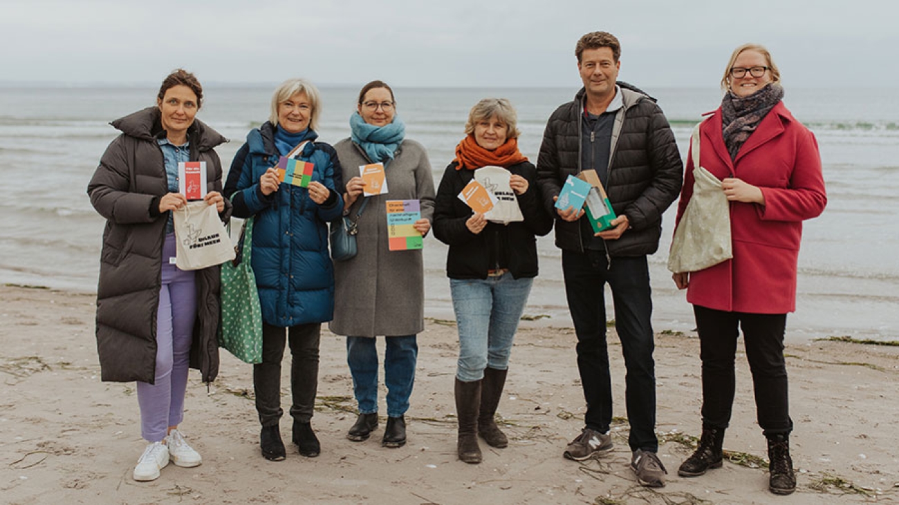 An der Ostsee stehen winterlich gekleidet fünf Frauen und ein Mann und halten Stoffbeutel und Prospekte in der Hand