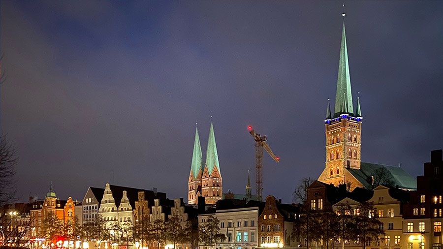 Ein abendliches Foto der beleuchteten Stadtsilhouette von Lübeck mit den Türmen von St. Marien und St. Petri