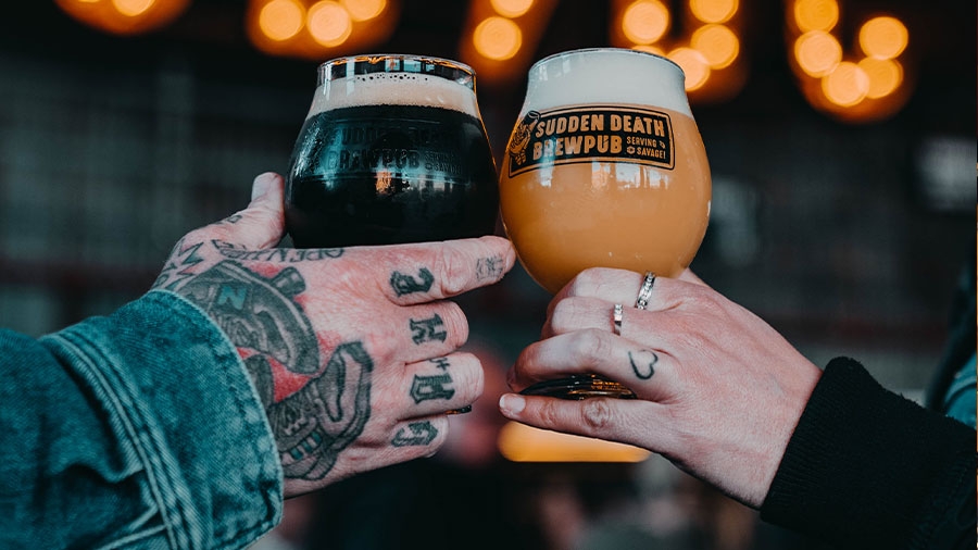 Zwei Hände stoßen mit Biergläsern an, auf denen "Sudden Death Brewpub" steht. Ein Glas enthält schwarzes Bier, ein anderes orange-milchiges